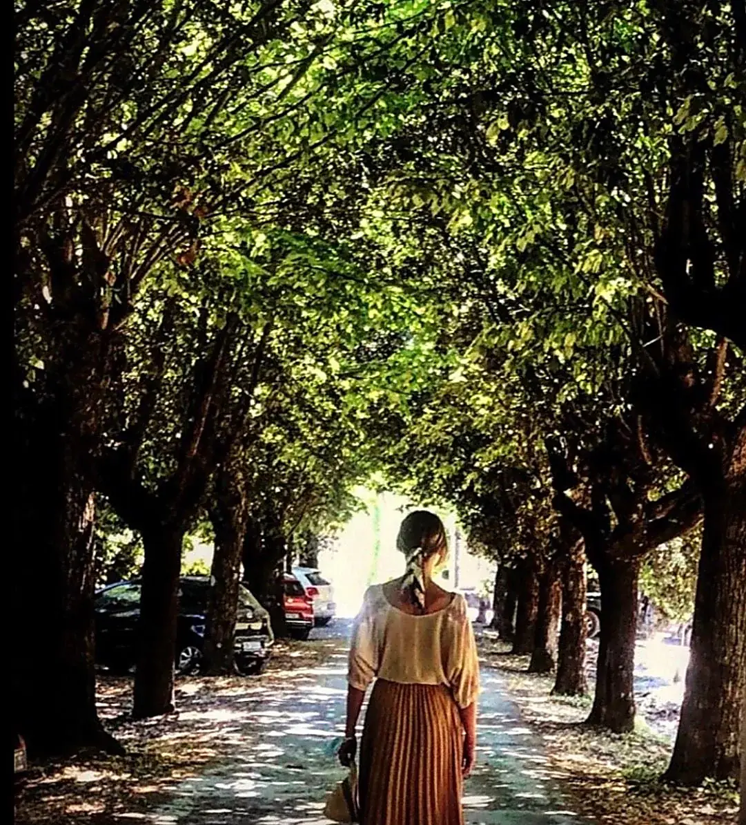A woman seen from behind walking along a shaded avenue of trees in the Italian countryside